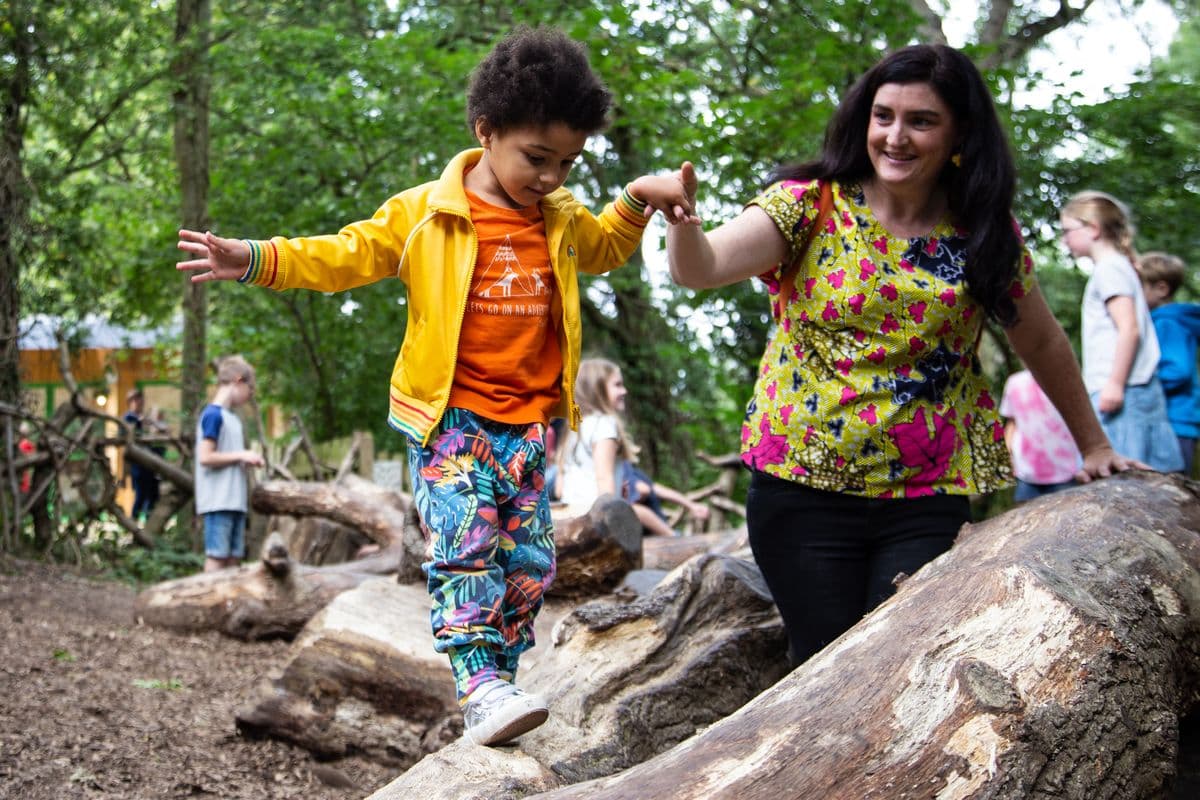 A young boy in a bright yellow raincoat climbs over some fallen tree branches whilst being supported by a smiling woman.