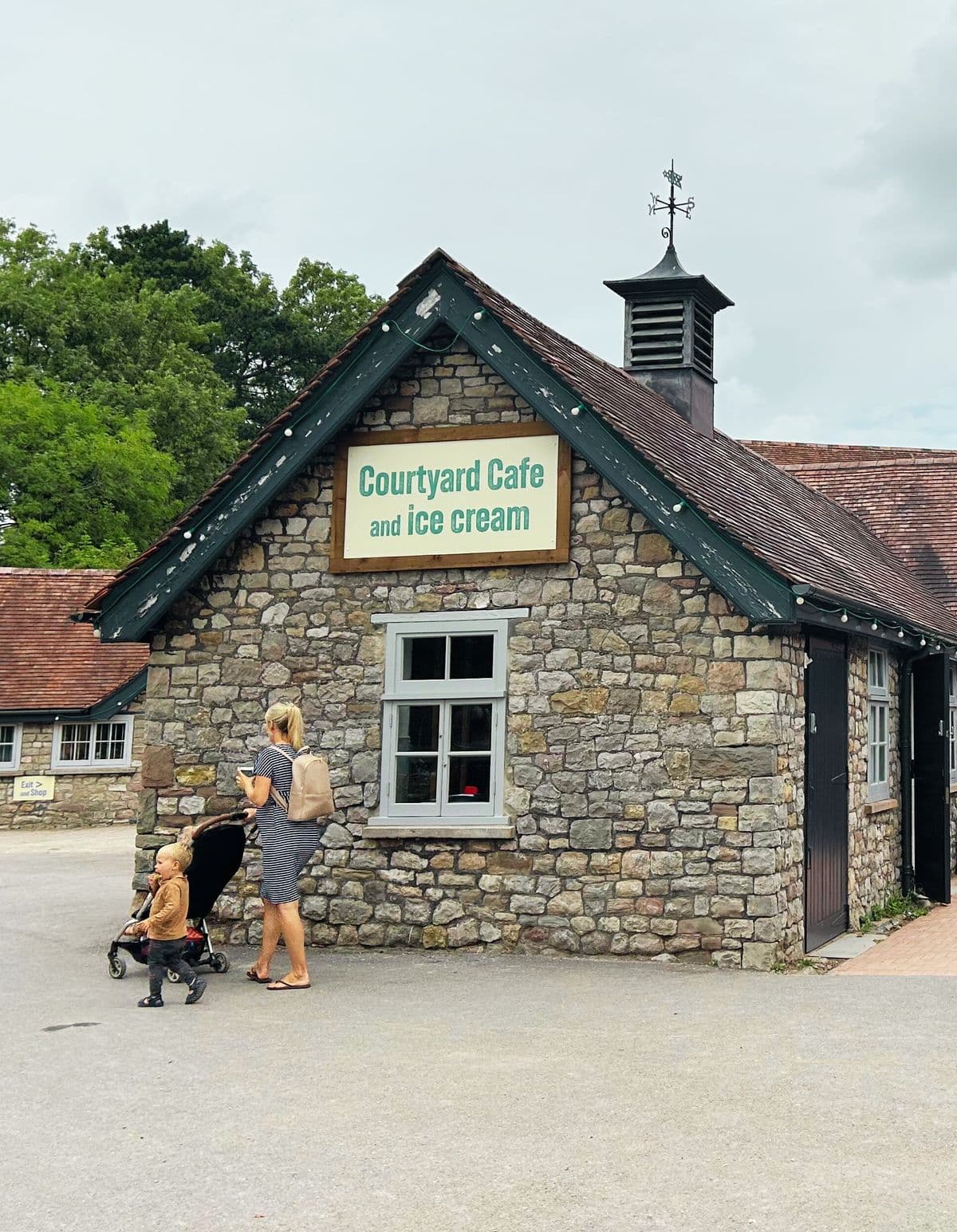 A woman pushing a pram holding a coffee walking past a stone café building