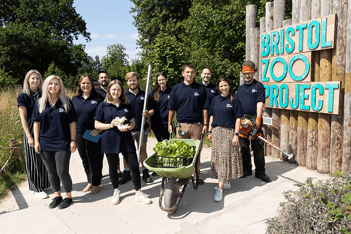 Eleven members of staff wearing their uniforms and holding props related to their jobs; standing in front of the Bristol Zoo Project sign
