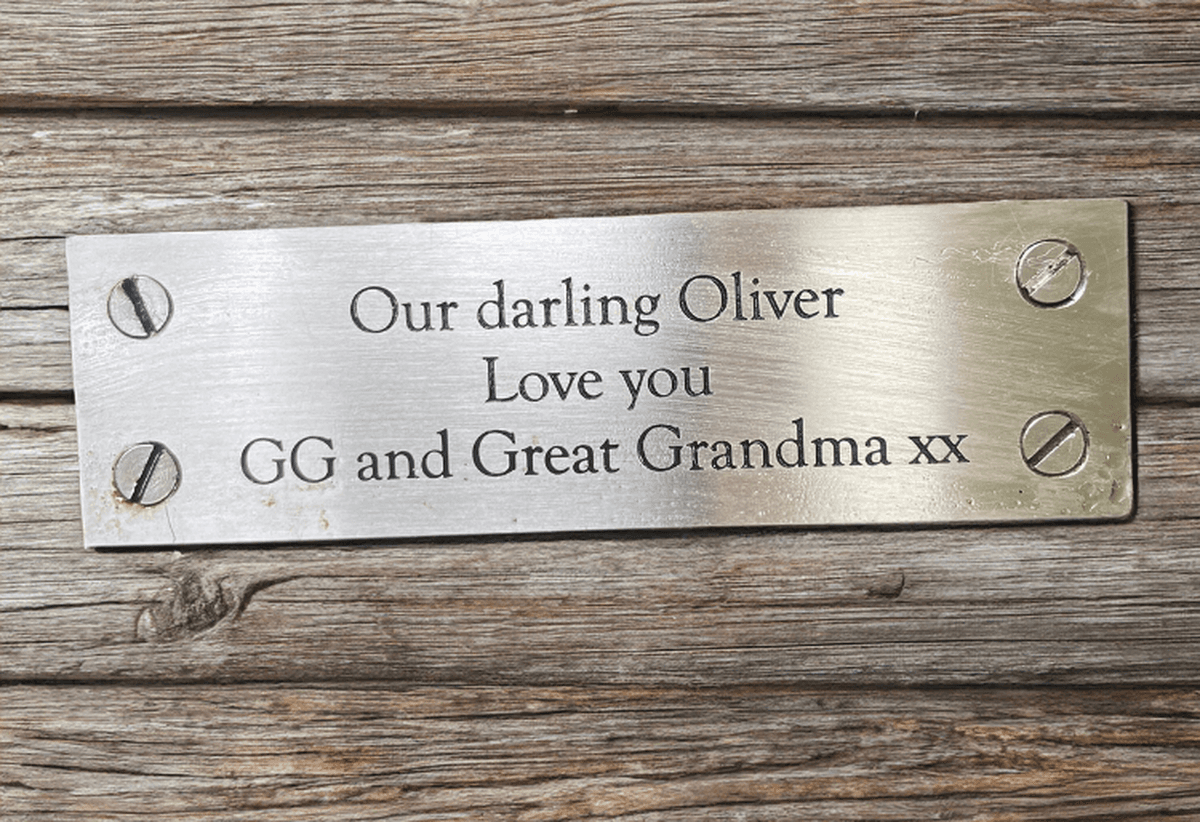 A gold-toned metal plaque fixed to a wooden walkway. The plaque has an engraved message which reads 'Our darling Oliver, Love you, GG and Great Grandma'