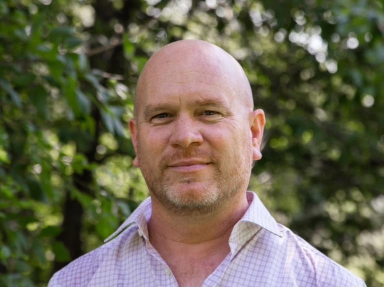 Brian is smiling in a close-up shot, he is wearing a lilac and white shirt.